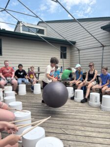Children attending a music therapy group. Each child plays an upside down bucket as a drum. A boy in the middle keeps beat with a ball.