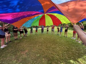 A group of teens hold a colorful parachute overhead.